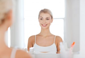 beauty, hygiene, morning and people concept - smiling young woman looking to mirror at home bathroom